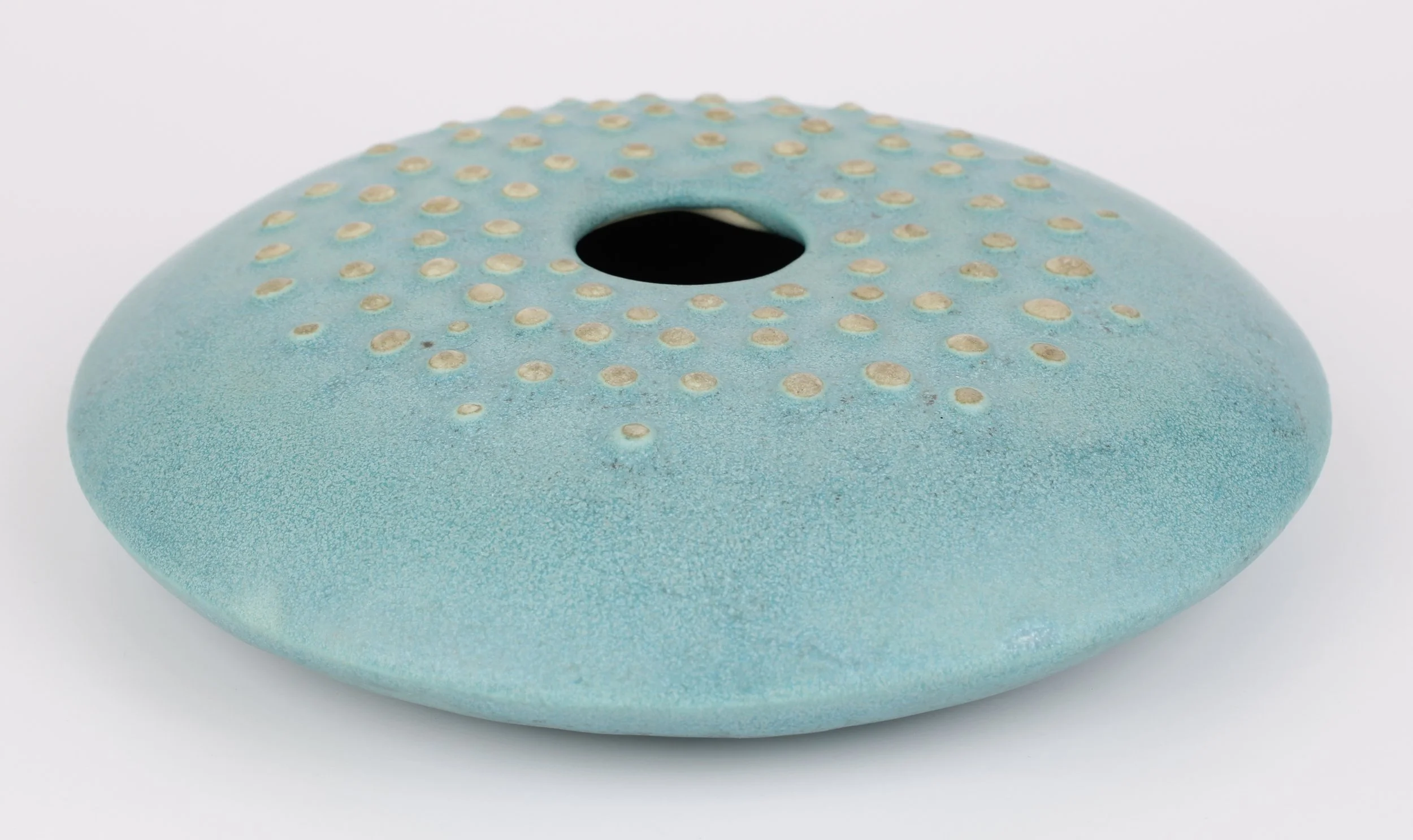 A round, blue ceramic object with a hole in the center and small beige dots on the top surface, resembling a donut.
