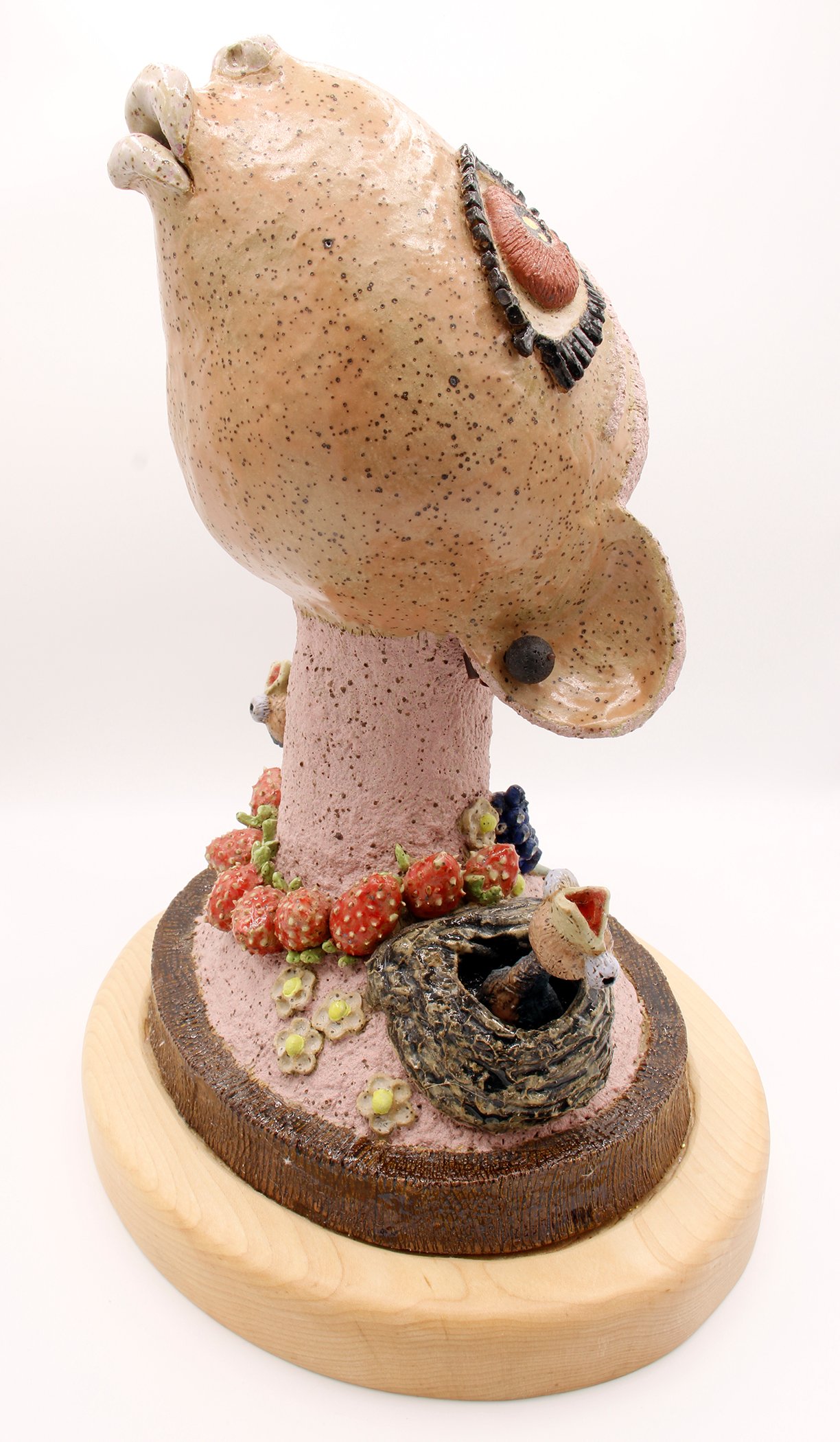A sculpture resembling a mushroom with various small objects attached, including strawberries, berries, and a bird in a nest, on a wooden base.
