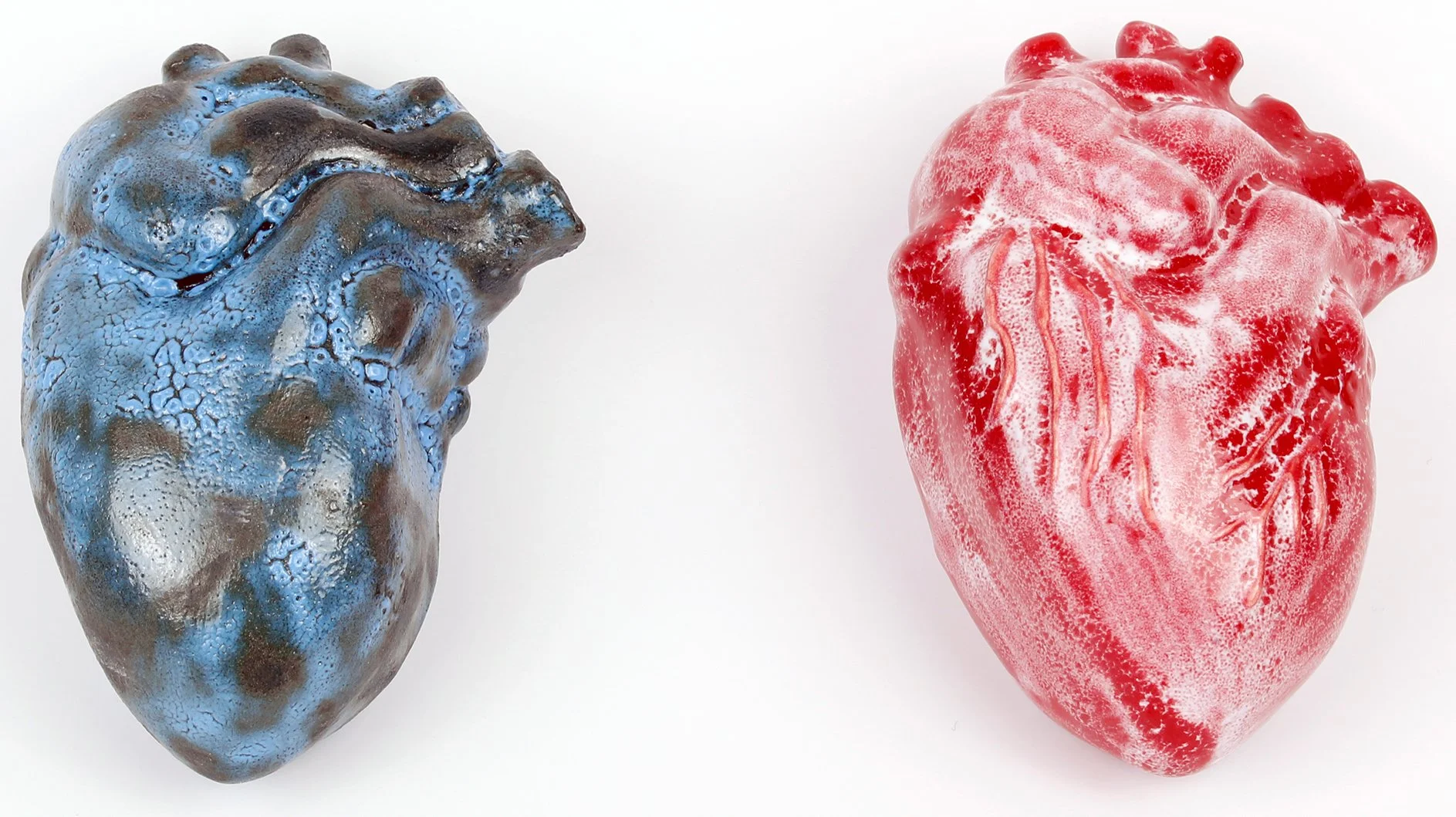 Two ceramic sculpture hearts, one painted in blue and black with a textured surface on the left, and the other painted in red with a marbled pattern on the right, against a white background.
