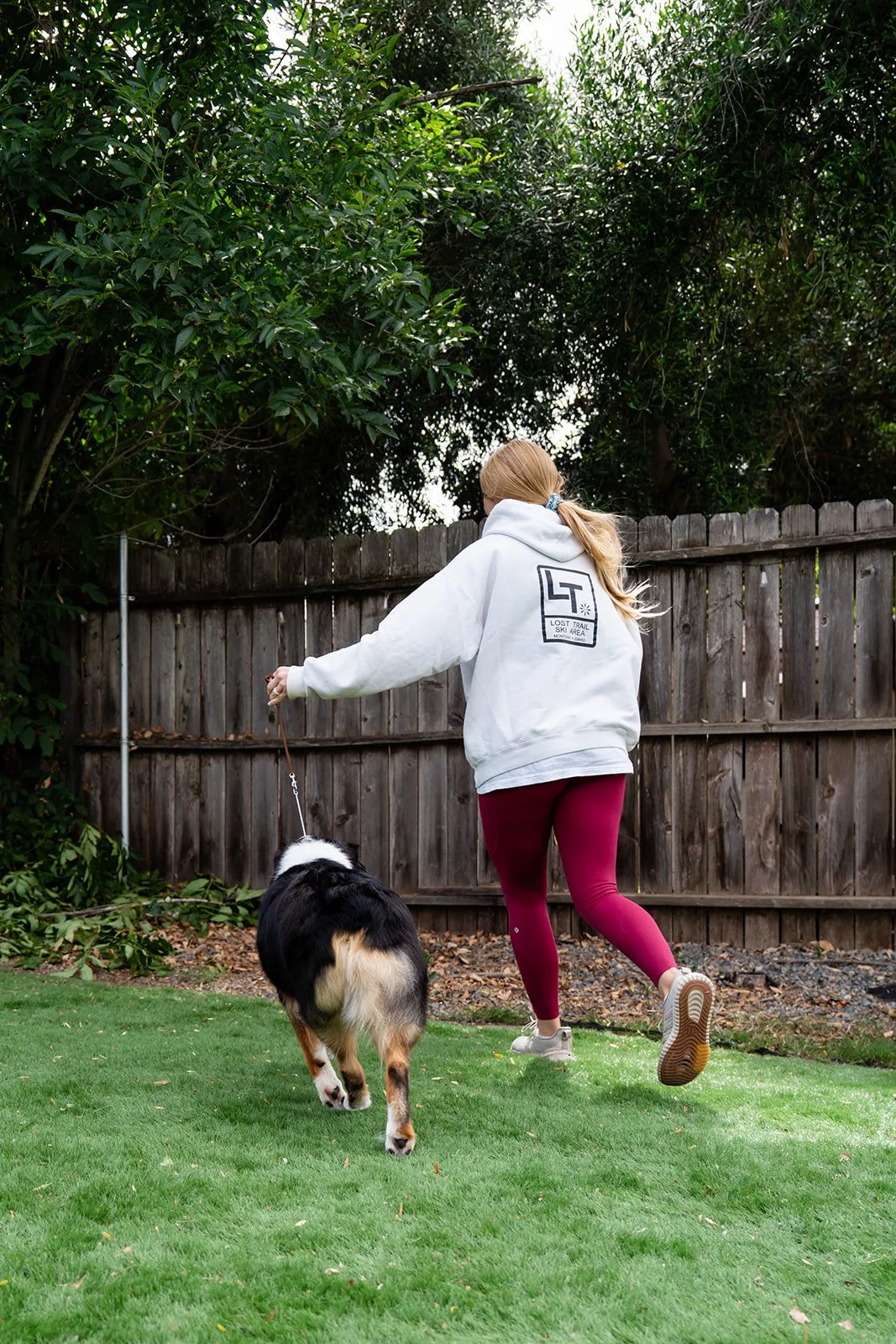 A woman walking a dog in a backyard with a wooden fence and green trees. Australian Shepherd breeder near me. Australian Shepherd puppy for sale.