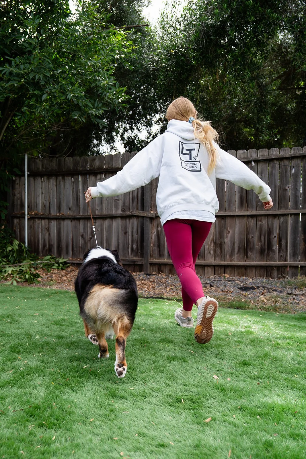 A woman in a white hoodie and maroon leggings runs on a green lawn while holding a leash attached to a black and white dog.
