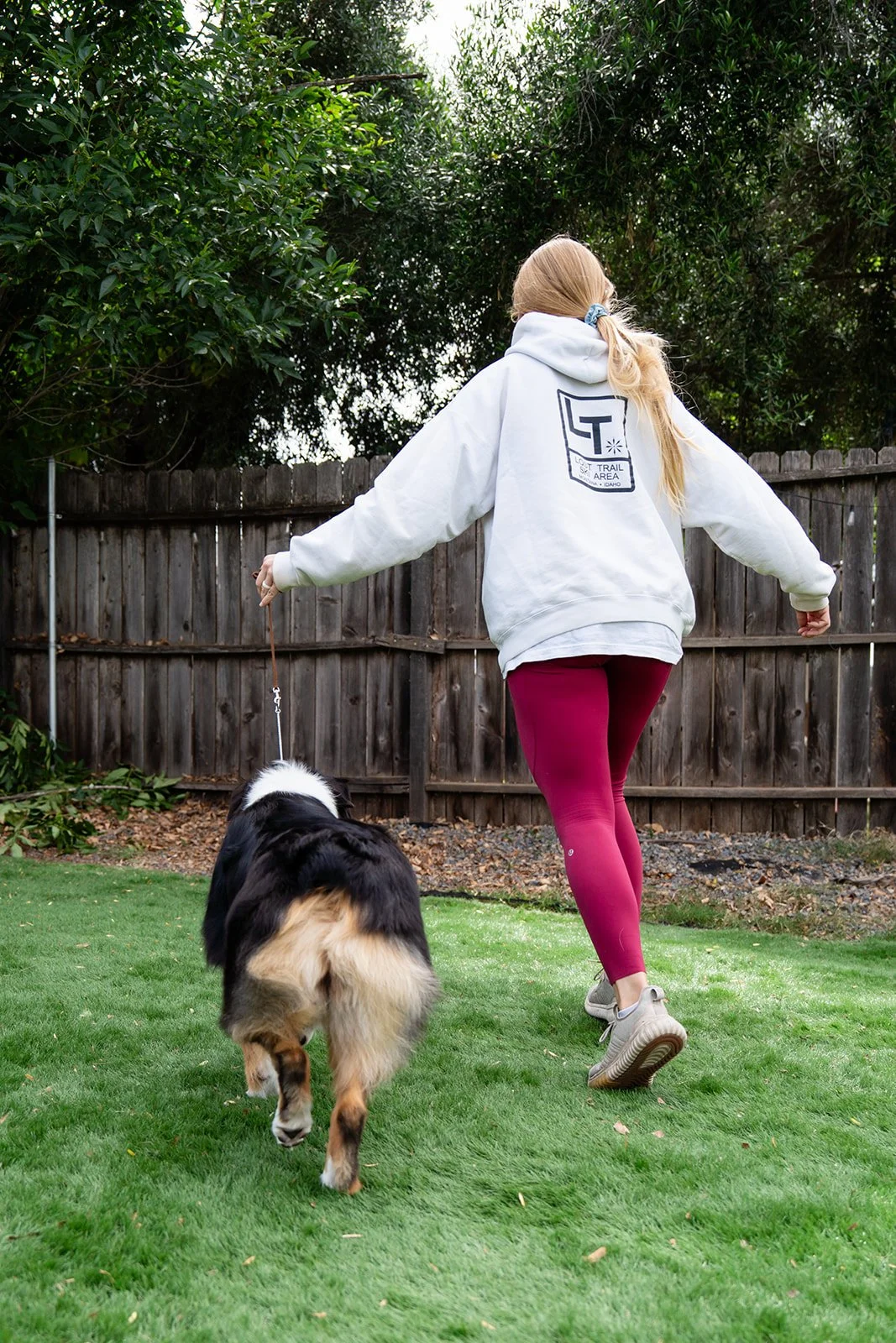 A woman in a white hoodie and maroon leggings walks a dog on a leash in a backyard with green grass and a wooden fence.