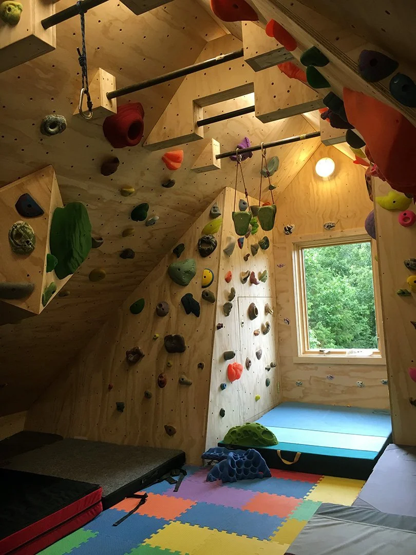 Indoor climbing gym with wooden walls, various colored climbing holds, and padded flooring.