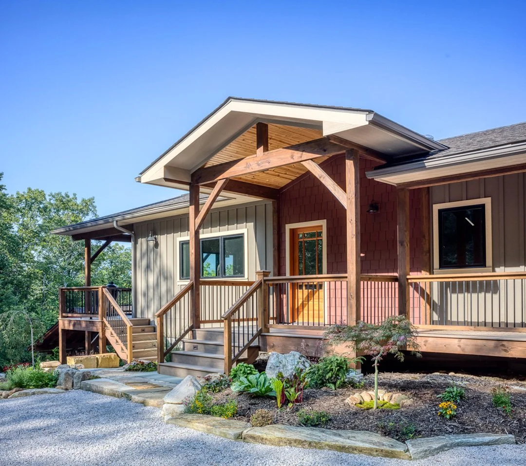 A modern house with a front porch, wooden railings, and steps, surrounded by a landscaped yard with plants and rocks.
