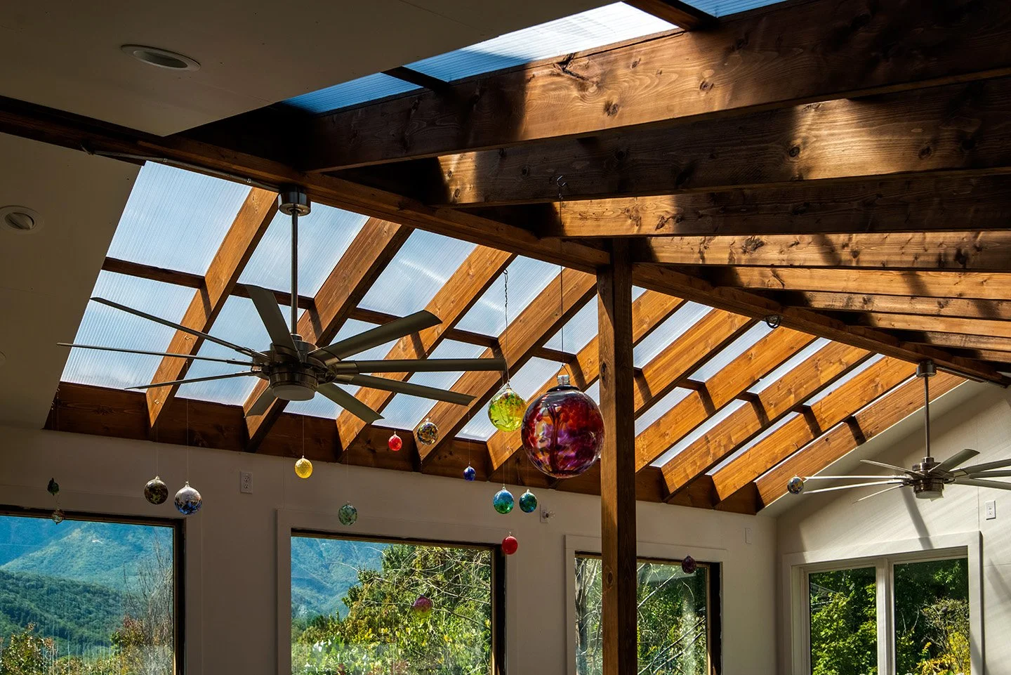 Interior view of a room with a wooden vaulted ceiling featuring skylights and a ceiling fan. Hanging colorful glass ornaments are suspended from the ceiling. Large windows show a scenic outdoor landscape with trees and mountains.