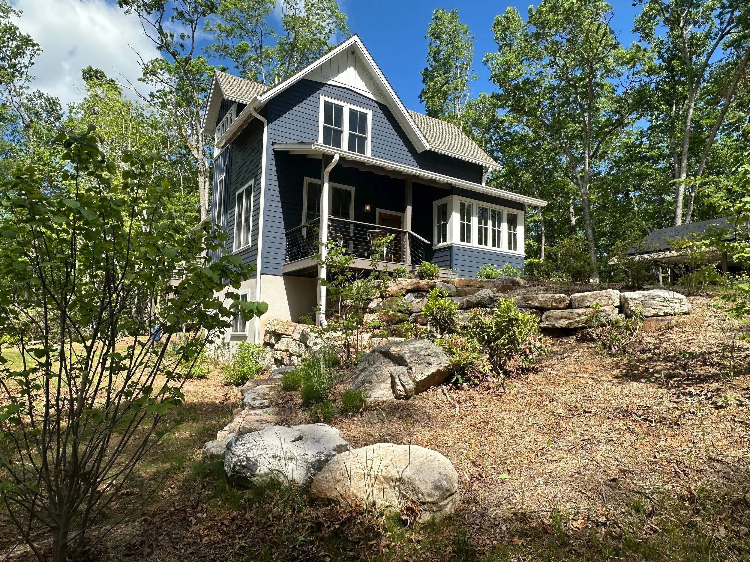 A blue two-story house with white trim, a small front porch, and a large bay window, situated on a sloped yard with rocks and plants, surrounded by trees under a partly cloudy sky.
