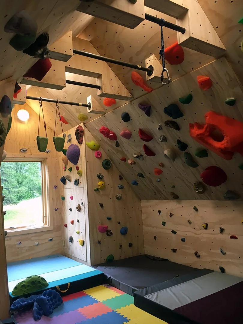 Indoor climbing gym with wooden walls, colorful climbing holds, mats on the floor, and a window showing greenery outside.