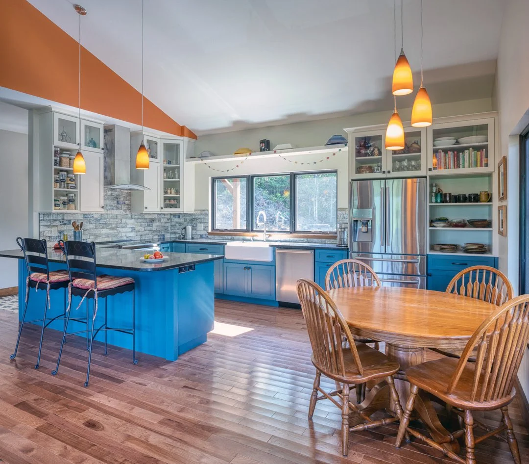 Modern kitchen with blue cabinets, white upper cabinets, kitchen island with two bar stools, round wooden dining table with six chairs, stainless steel refrigerator, pendant lights, and hardwood flooring.