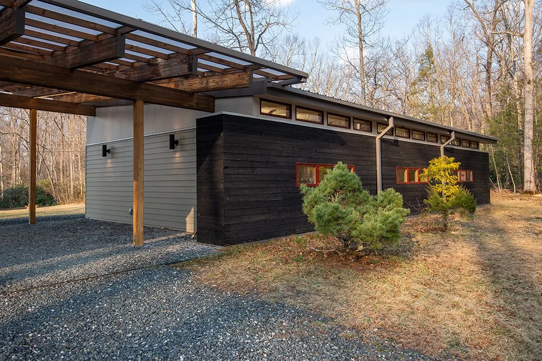 Modern house with dark and light siding, small horizontal windows, surrounded by trees and bushes, with gravel driveway and an overhanging wooden roof structure.