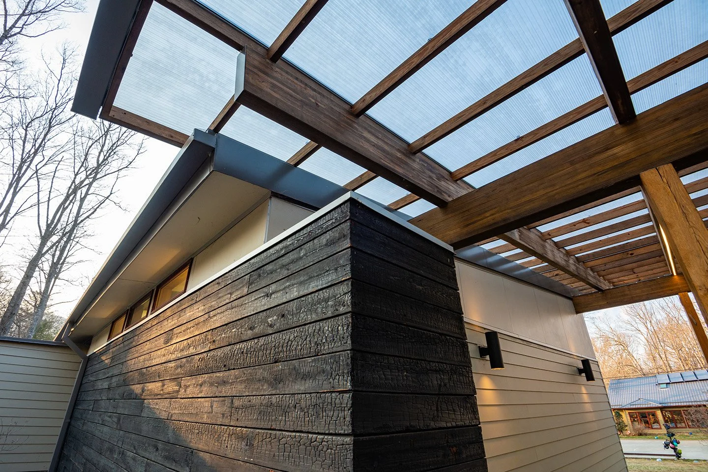 Modern house with black wooden exterior siding and a transparent roof with wooden beams, outdoors with trees and other buildings in the background.
