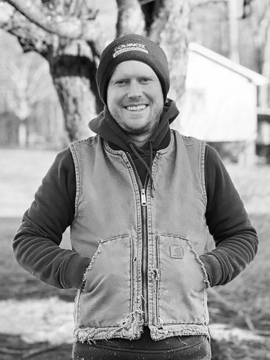 A smiling man standing outdoors in a black and white photo, wearing a beanie, hoodie, and a sleeveless jacket.