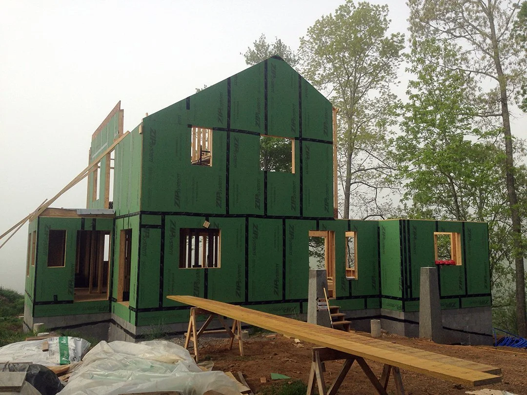 Construction site with a partially built two-story house with green sheathing panels, wooden framing, and a ramp leading up to the entrance in a wooded area.