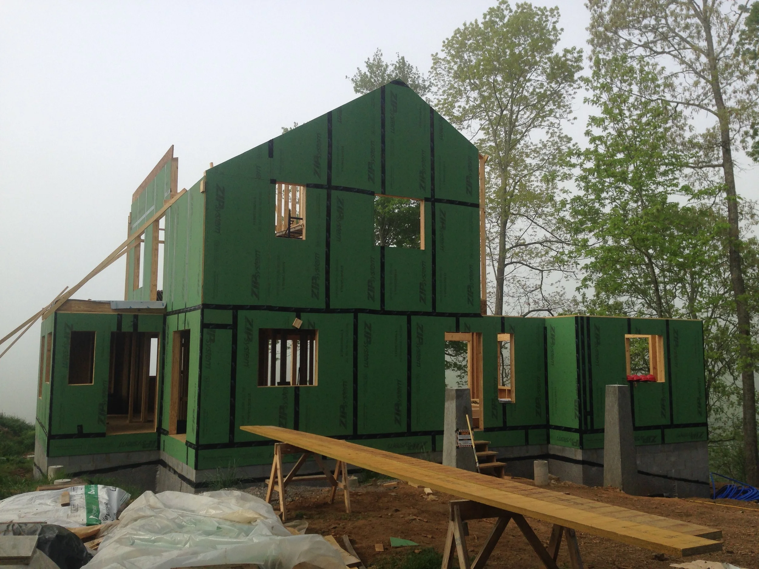 Construction of a two-story house with green wall sheathing, wooden framing, and window openings, surrounded by trees and construction materials.