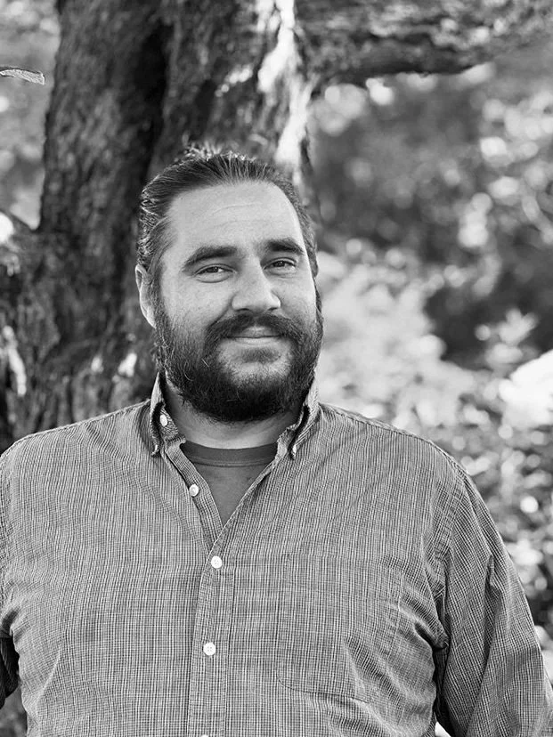 A man with a beard and slicked-back hair standing outdoors next to a tree, wearing a checked button-up shirt and smiling.