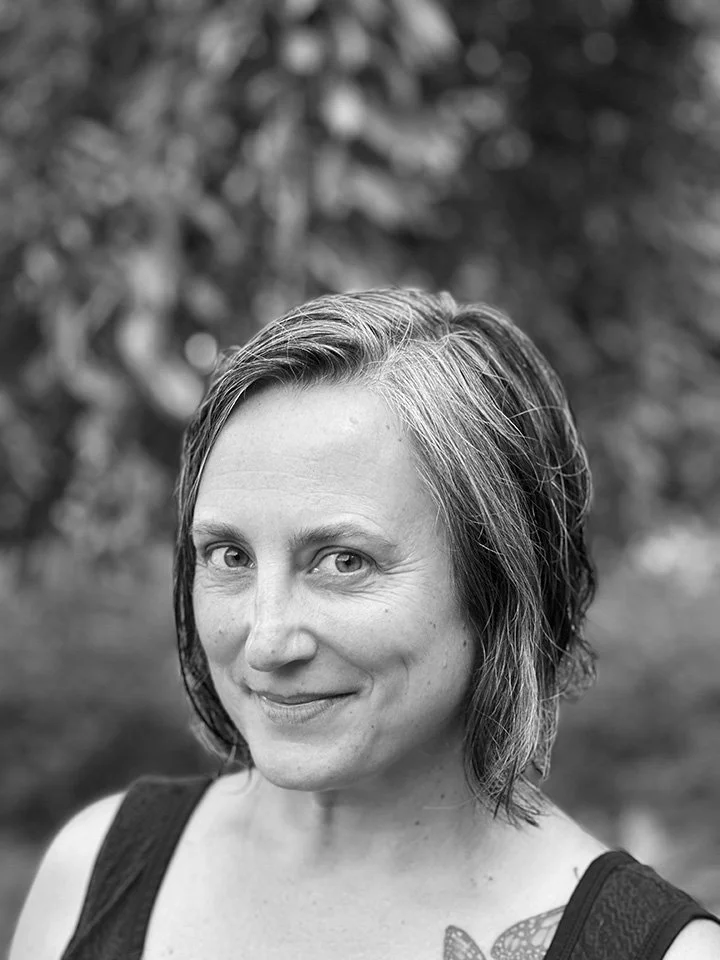 Black and white photo of a woman with short, wavy hair, smiling slightly, outdoors with a blurred background of trees.