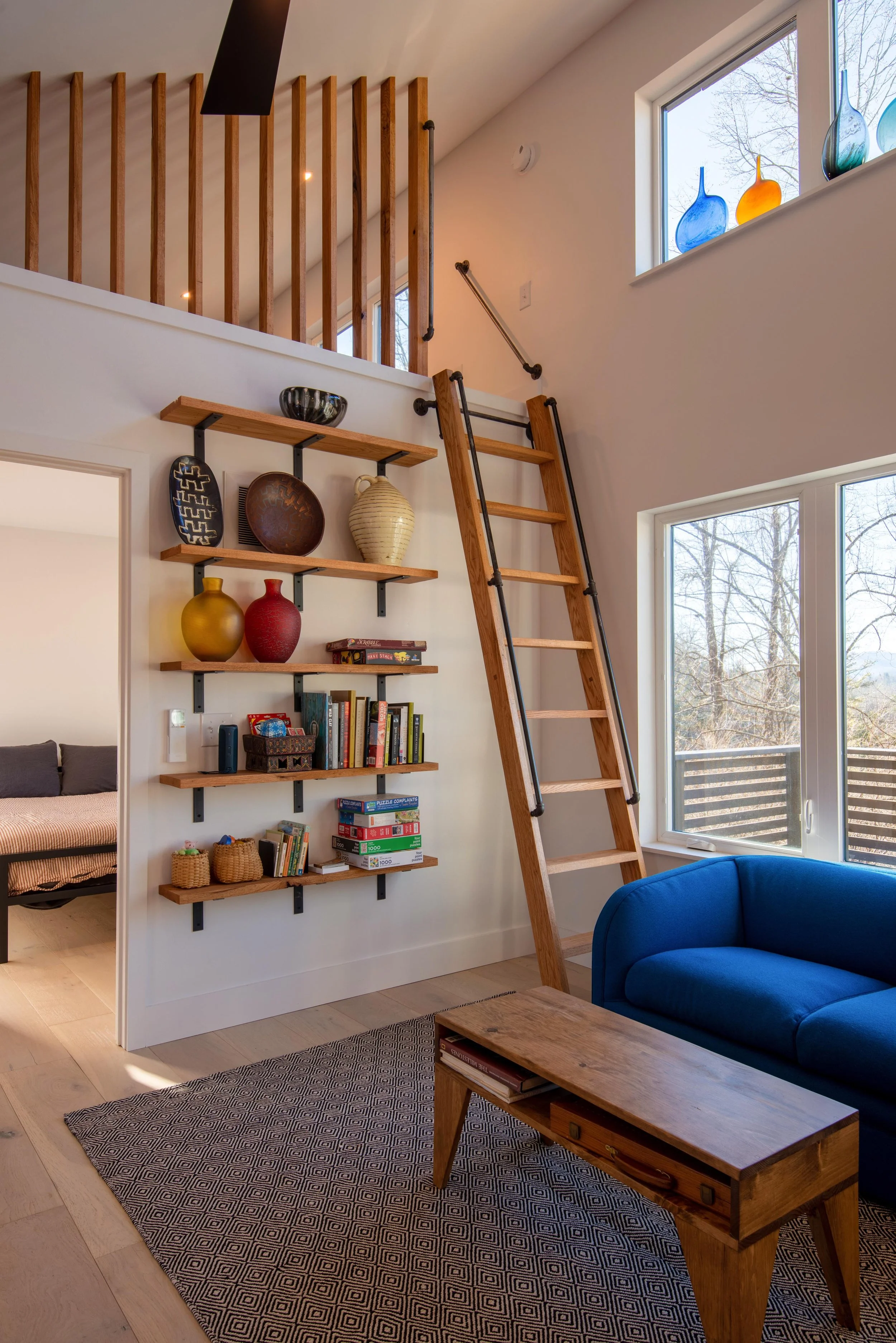 Interior of a living room featuring a wooden ladder, a large window with colored glass vases on the sill, a blue couch, a wooden coffee table, and a wall-mounted shelf with decorative items and books.