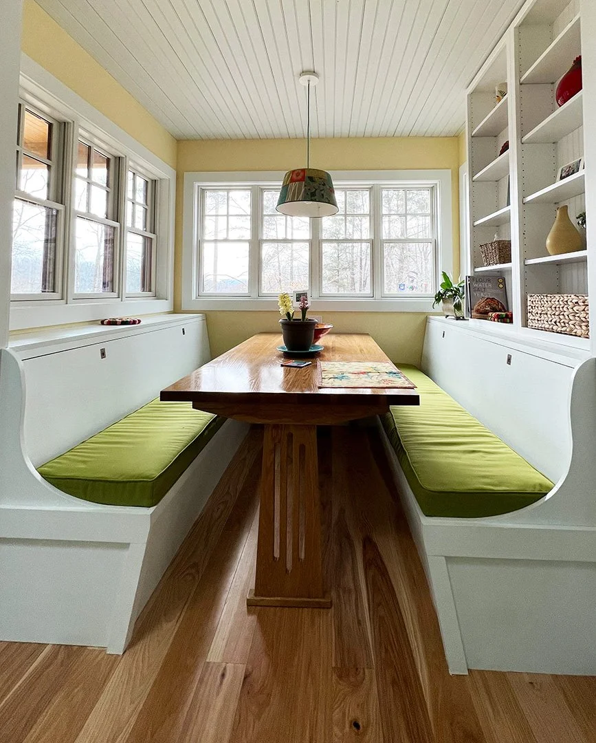 A sunlit kitchen nook with a wooden table, built-in white bench seats with green cushions, large windows, and open white shelving on the right side, decorated with dishes and plants.