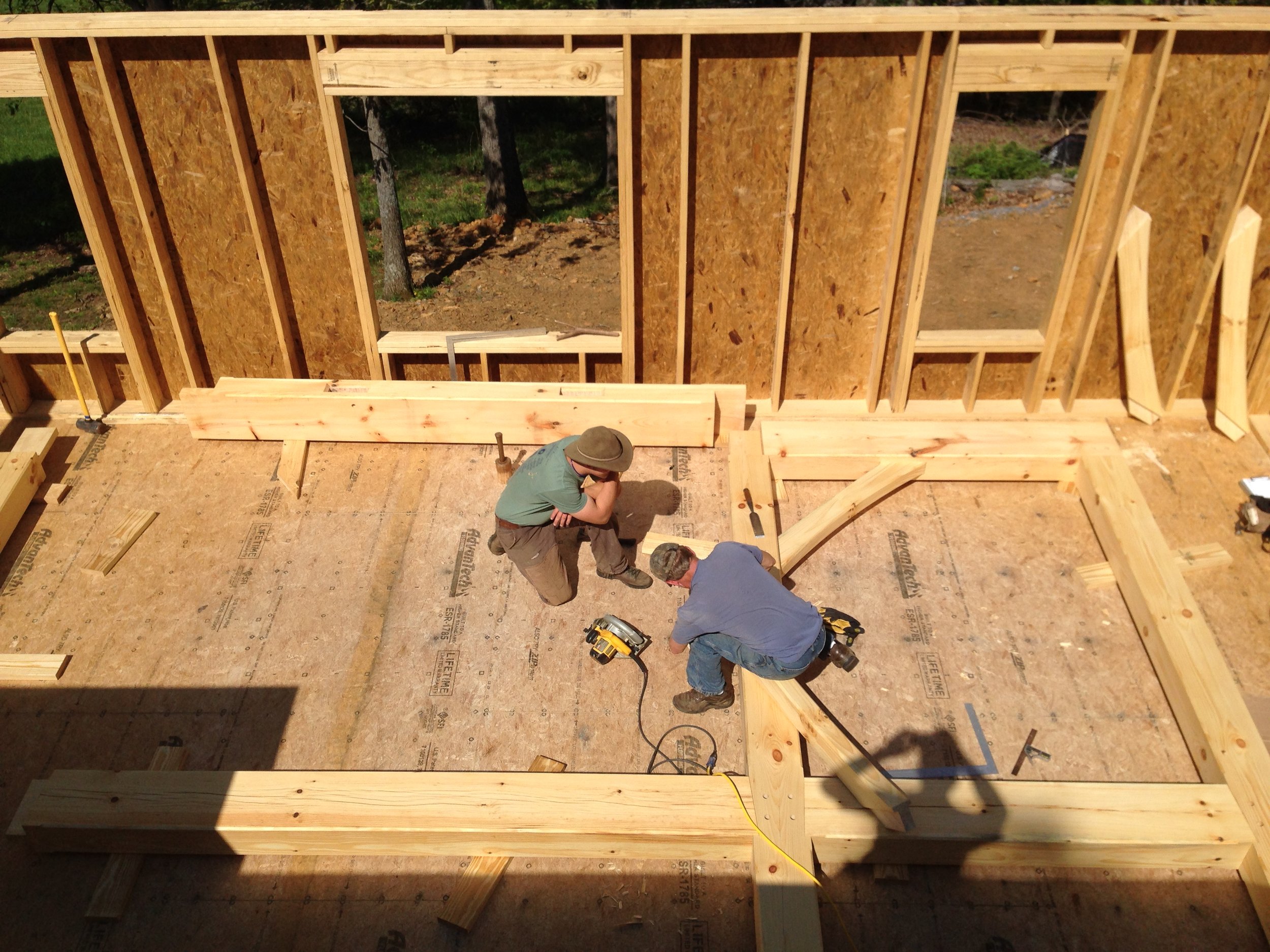 Two construction workers install wooden beams on the floor of a house under construction, with unfinished wooden walls and open window framing behind them.