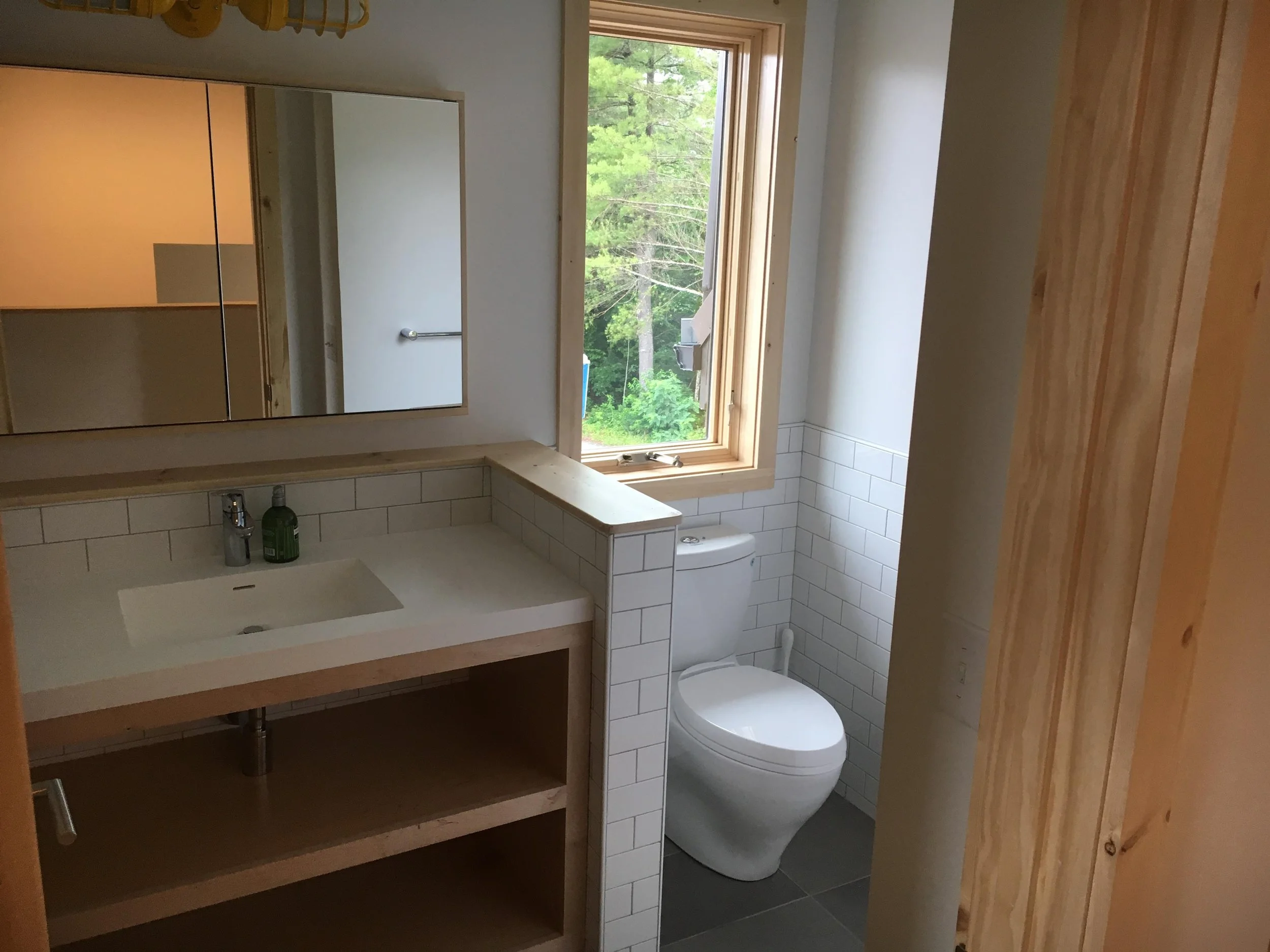 A bathroom with a wooden vanity, a mirror, a white sink, a soap dispenser, a window showing greenery outside, and a modern white toilet.
