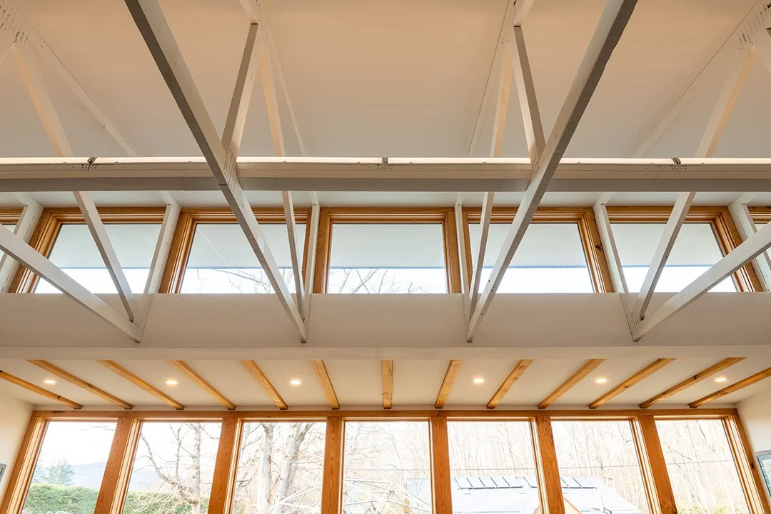 Interior view of a modern house with large windows, some with wooden frames, and a high ceiling with exposed wooden beams. The ceiling is under construction with visible framework and incomplete plaster or drywall.