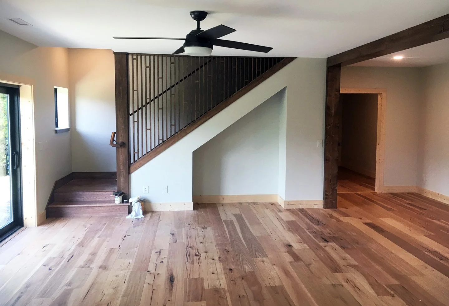 Interior of a house with wooden floors, staircase with a rustic wooden post and black railing, ceiling fan, and doorway leading to another room.
