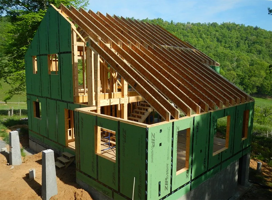 A house under construction showing the wooden framing and green exterior sheathing, with a steeply sloped roof and multiple window openings, set in a green outdoor area.