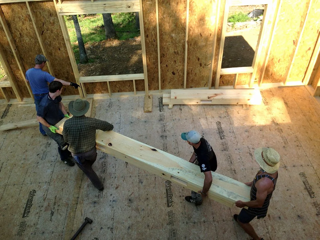 Five people working together to hold a large wooden beam inside a house under construction, with wooden wall studs and open window frames visible.