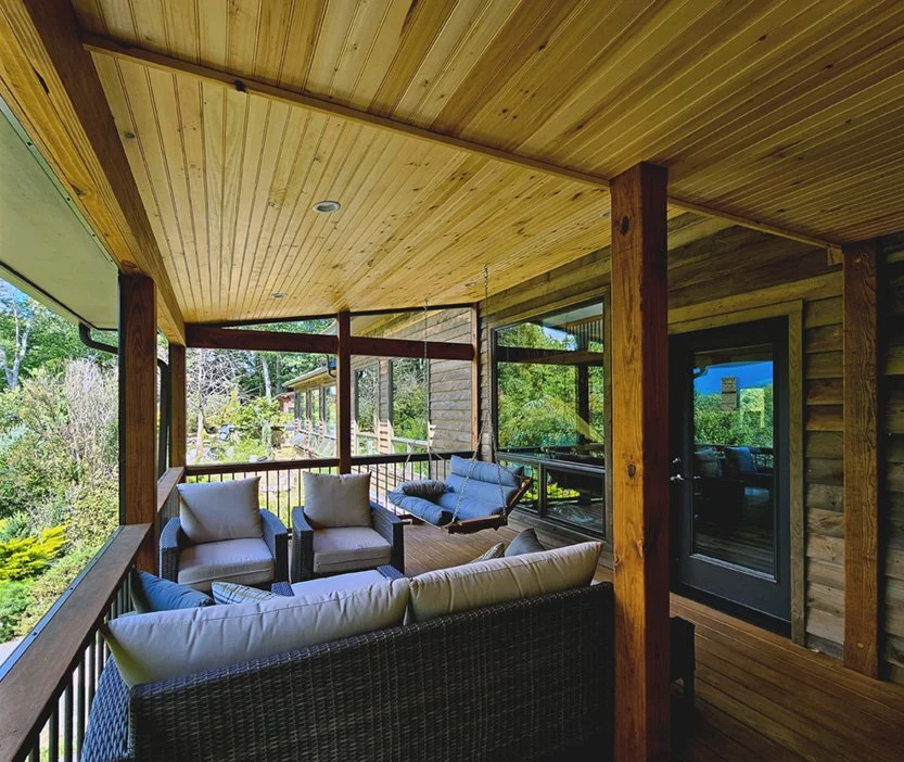 A screened-in porch with wooden ceiling and walls, featuring outdoor seating including a sofa, two armchairs, and a hanging swing, overlooking a lush green garden.