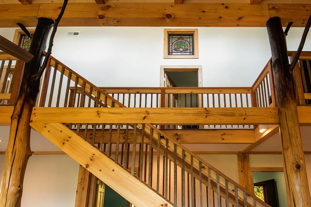 Interior view of a two-story wooden staircase with railing, wooden beams, and decorative stained glass window.