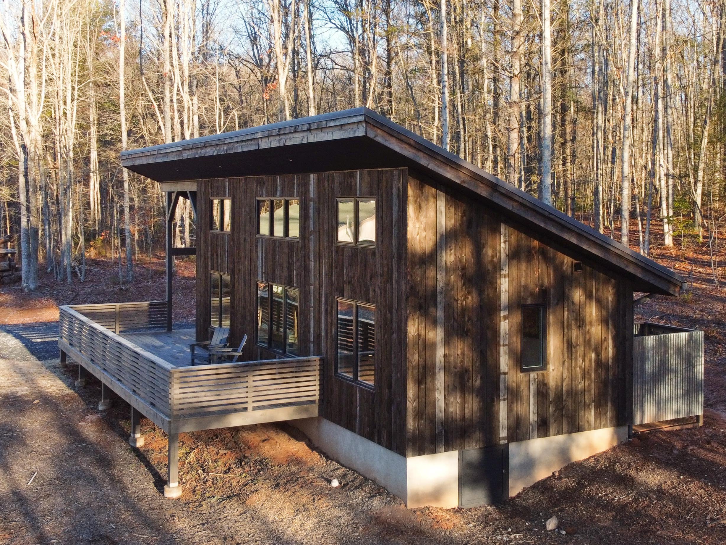 A modern two-story house with dark wood siding, multiple windows, and a sloped roof, situated in a wooded area with leafless trees.
