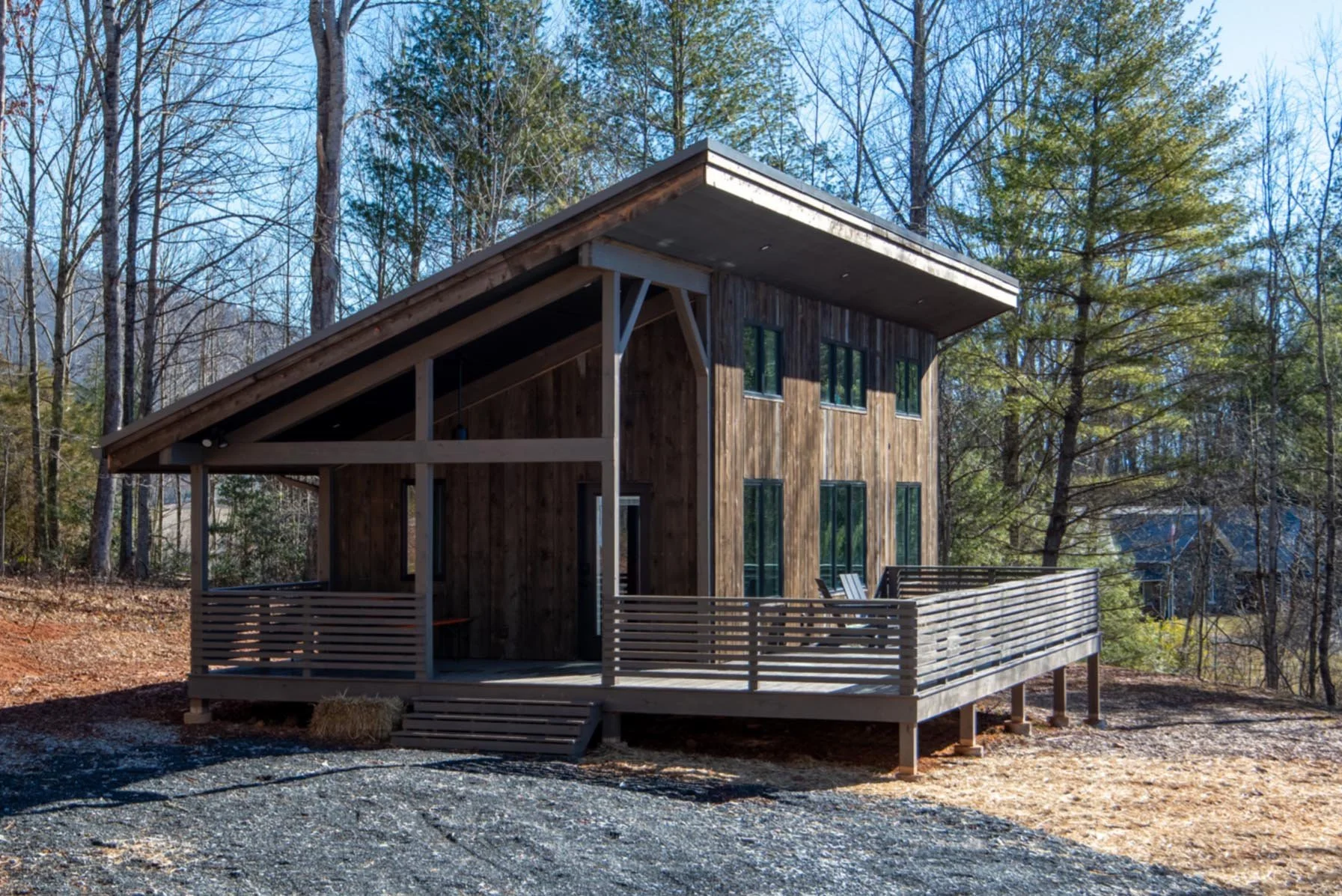 A modern two-story wooden house with a sloped roof, large windows, and a wraparound deck in a wooded area with trees.