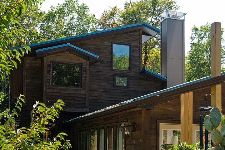 A two-story wooden house with large windows, a sloped roof, and a chimney, surrounded by trees and greenery.