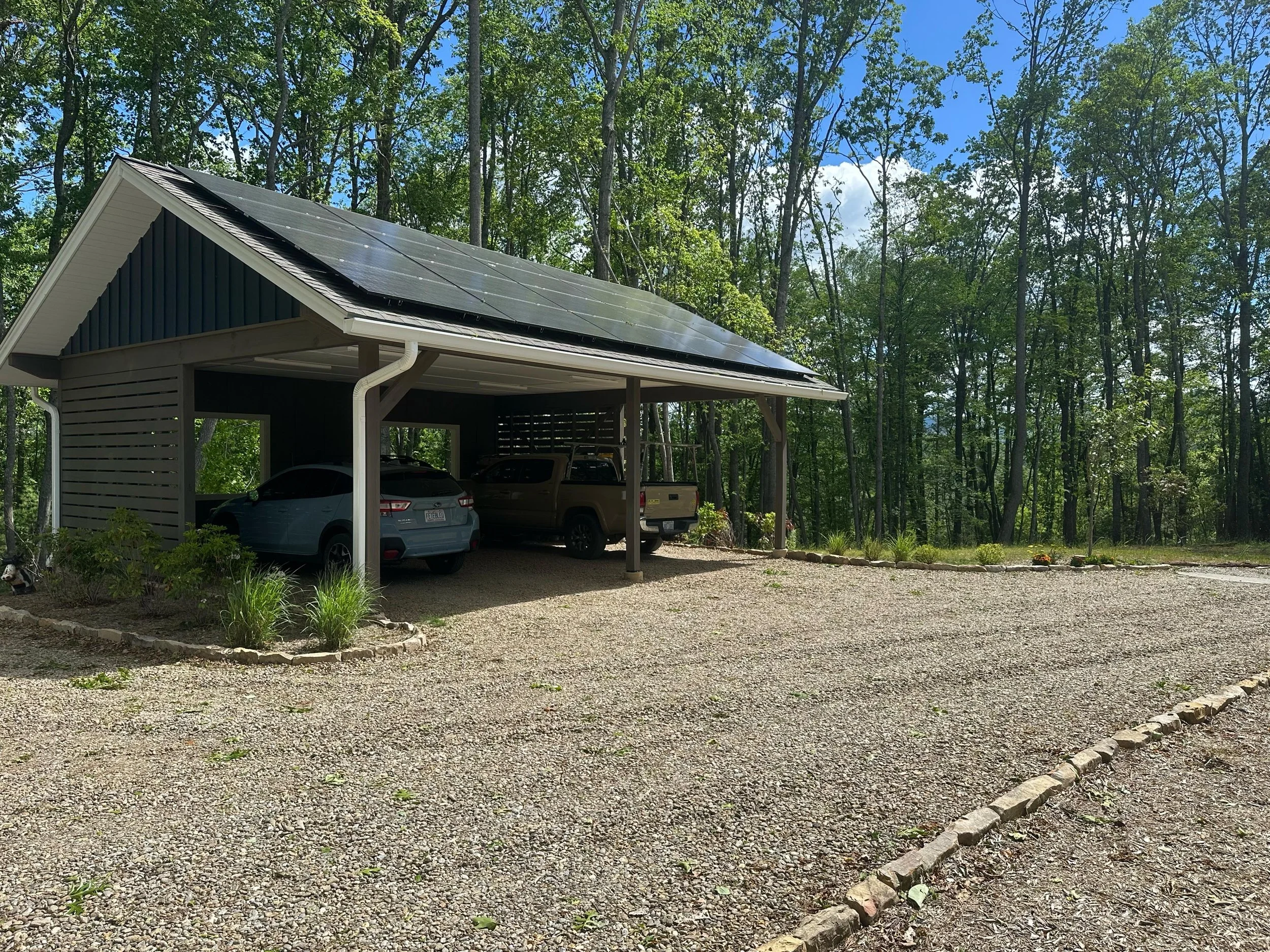 A carport with a gravel driveway, two vehicles parked underneath, and a wooded forest background.