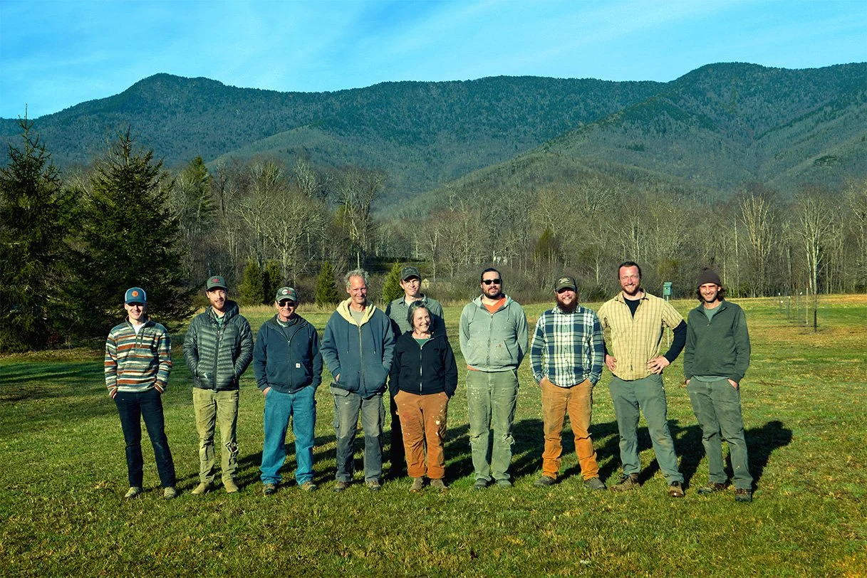 Group of ten people standing outdoors on grass with mountains and trees in the background.