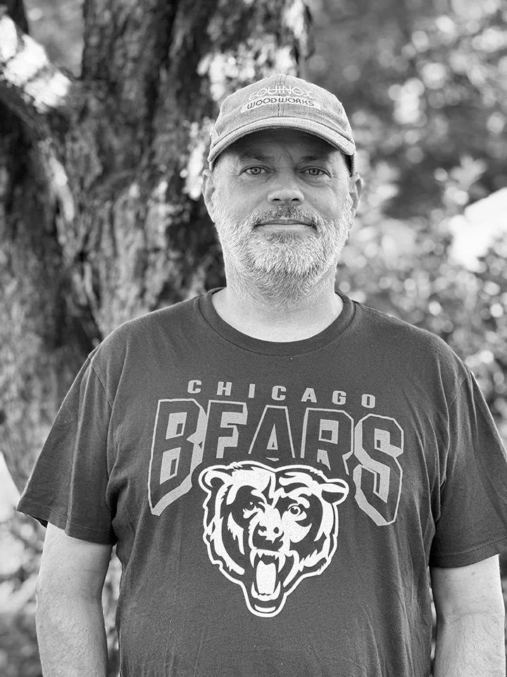 A man with a beard wearing a baseball cap and a Chicago Bears t-shirt, standing outdoors near a tree in black and white.