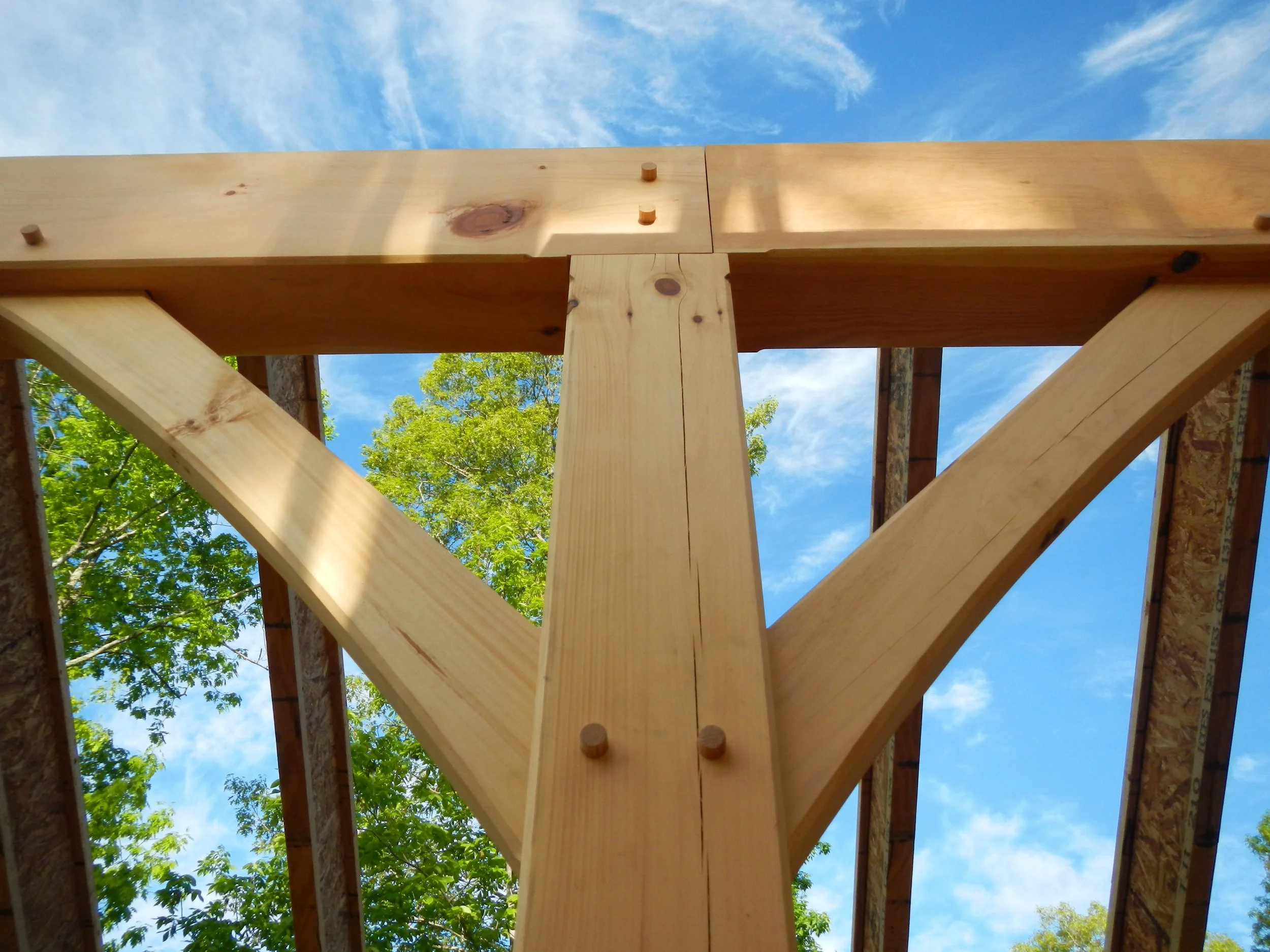 Close-up view of a wooden construction framework with beams and support structures under a blue sky with clouds and trees in the background.