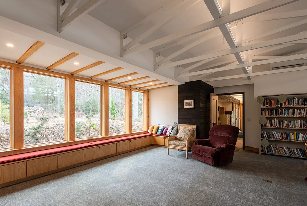 Bright living room with large window seat, featuring colorful pillows, facing a wooded outdoor scene. Includes a maroon armchair, a patterned armchair, and a bookshelf on the right.