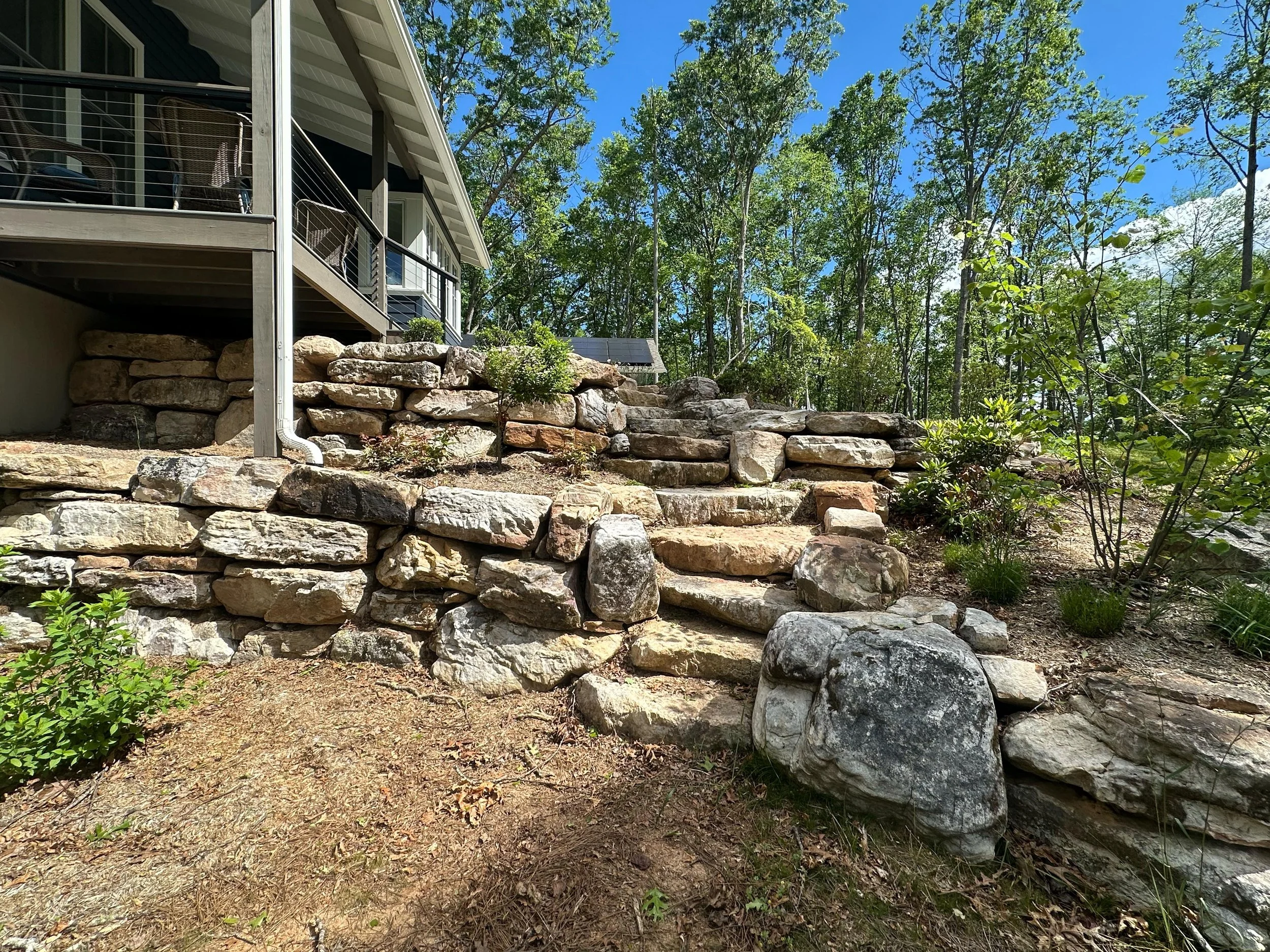 Stone staircase leading up a hill next to a house with a deck, surrounded by trees and greenery under a blue sky.