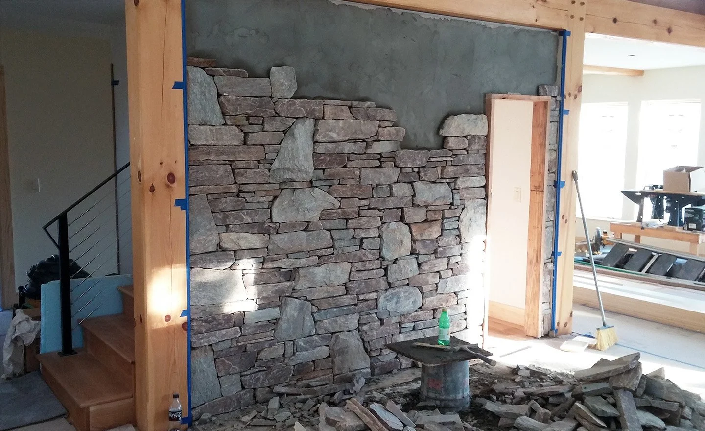 A stone wall under construction with large and small irregularly shaped rocks, in a room with wooden framing and construction tools nearby.