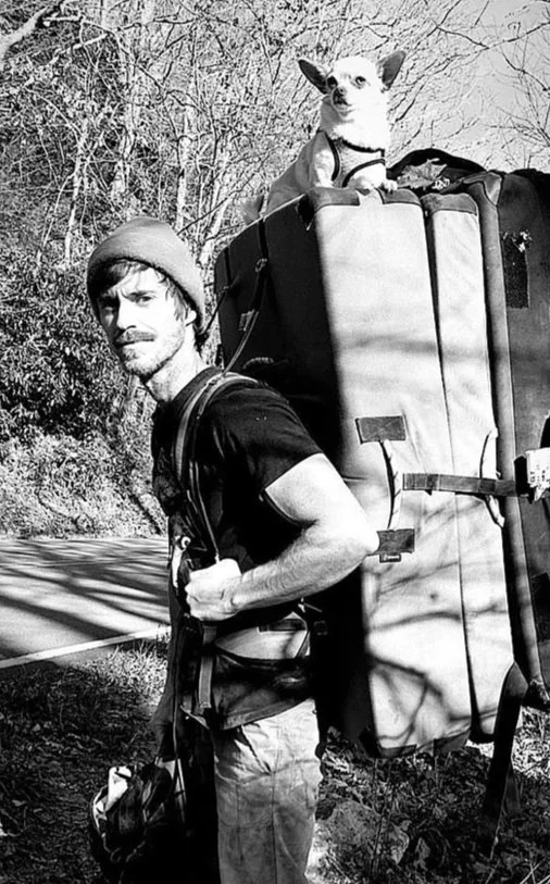A man hiking with a small dog sitting in a backpack carrier on his back, outdoors with trees and a road in the background.