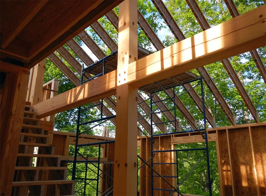 View of a wooden house under construction with scaffolding and tree-lined background.