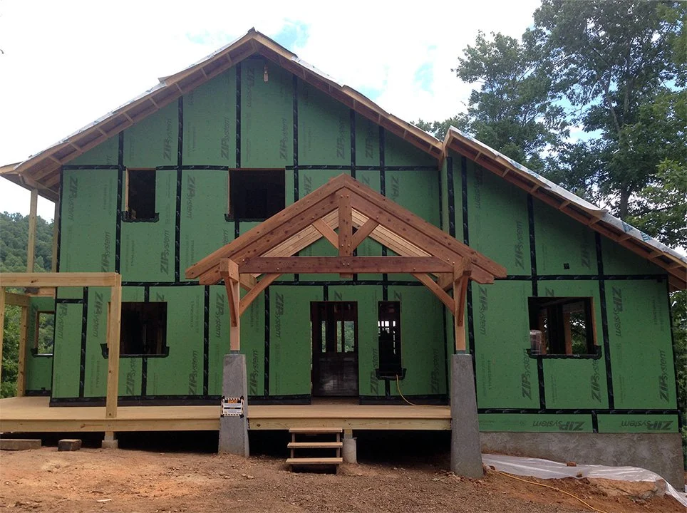 A house under construction with green sheathing and a wooden porch with a small gabled roof, surrounded by trees.