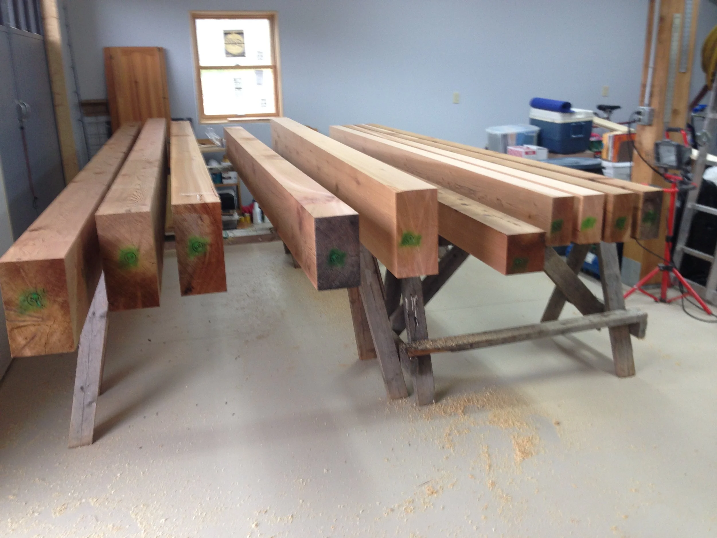 Workshop with several large wooden beams on sawhorses, inside a well-lit garage with tools and supplies in the background.