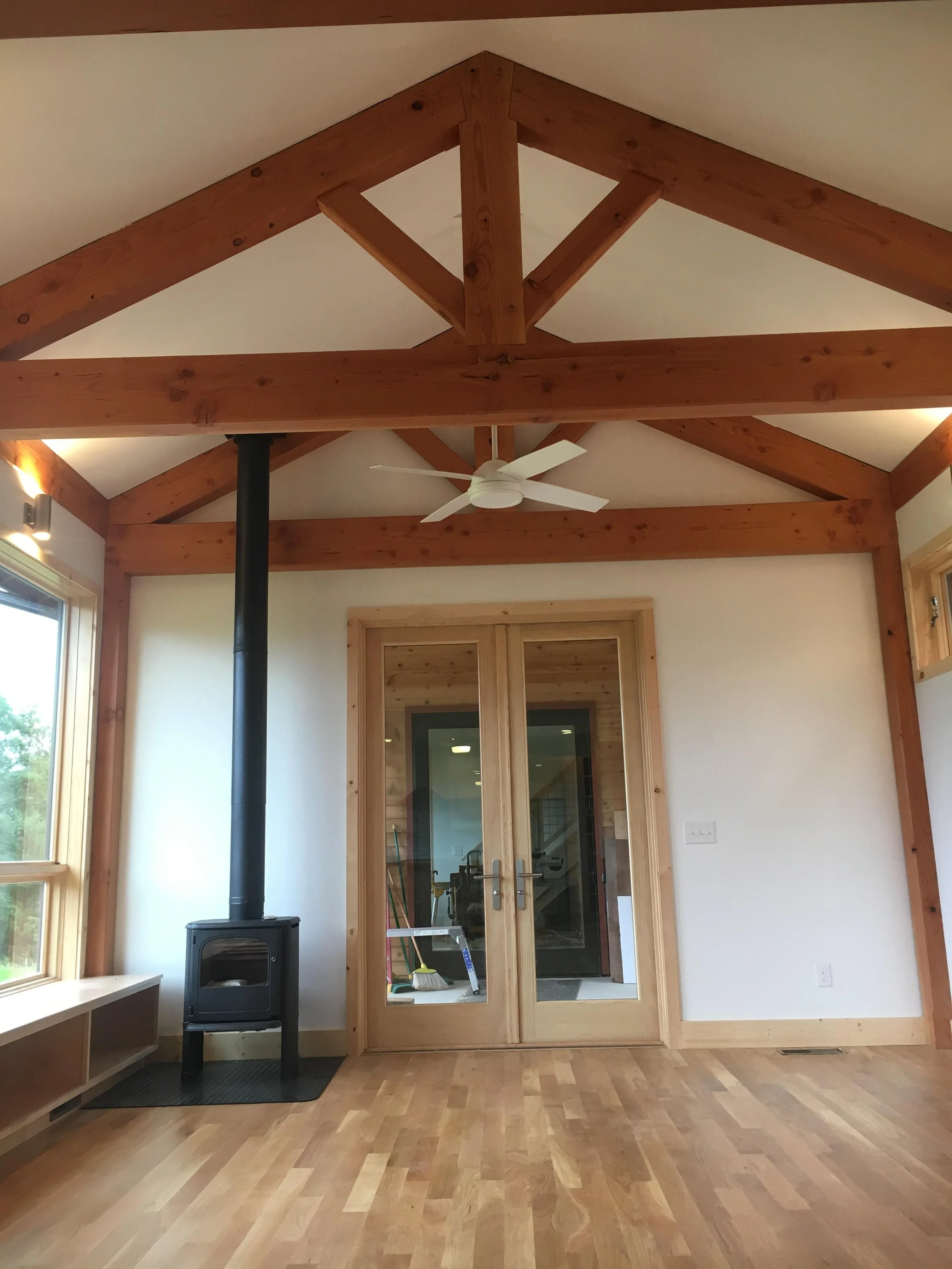 Room with wooden beams on ceiling, a ceiling fan, a wood stove with a chimney pipe, large windows, and a set of French doors leading to another room.