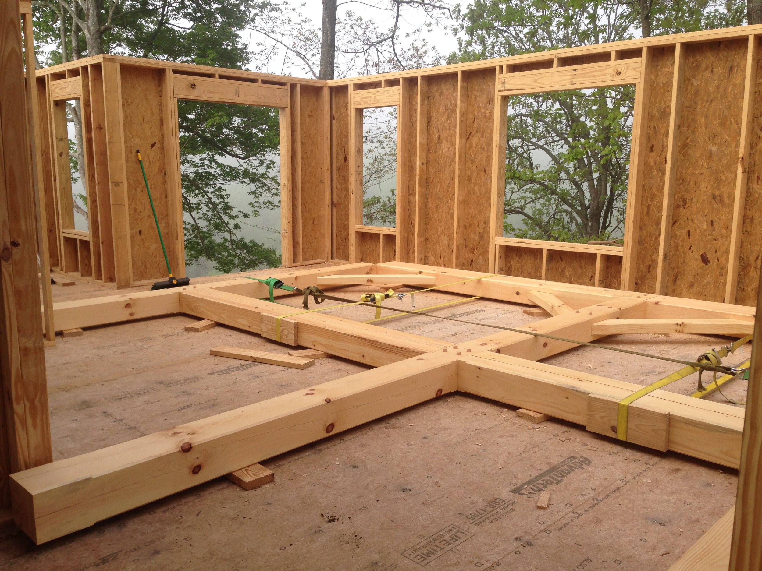 The image shows an unfinished wooden room under construction with walls and floor framing, large open windows, and some construction tools and materials.