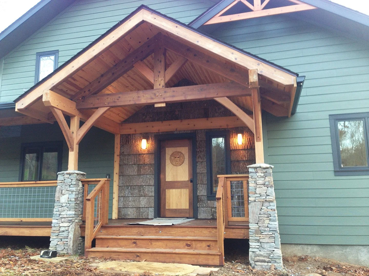 Front entrance of house with wooden porch, stone columns, green siding, and black framed windows.