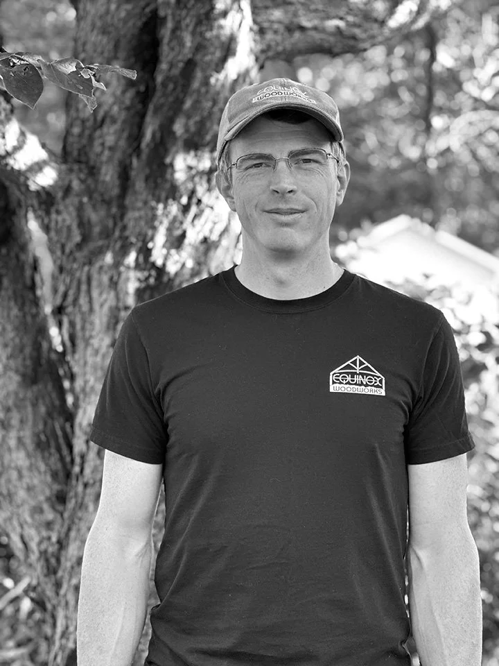 A man standing outdoors next to a tree, wearing glasses, a cap, and a dark t-shirt with a logo.