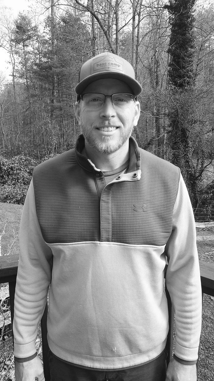 A man wearing glasses, a baseball cap, and a fleece jacket stands outdoors in a wooded area, smiling at the camera.