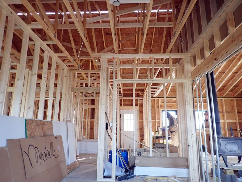 Interior of a house under construction with wooden framing, a carpenter working in the background, and construction materials scattered around.