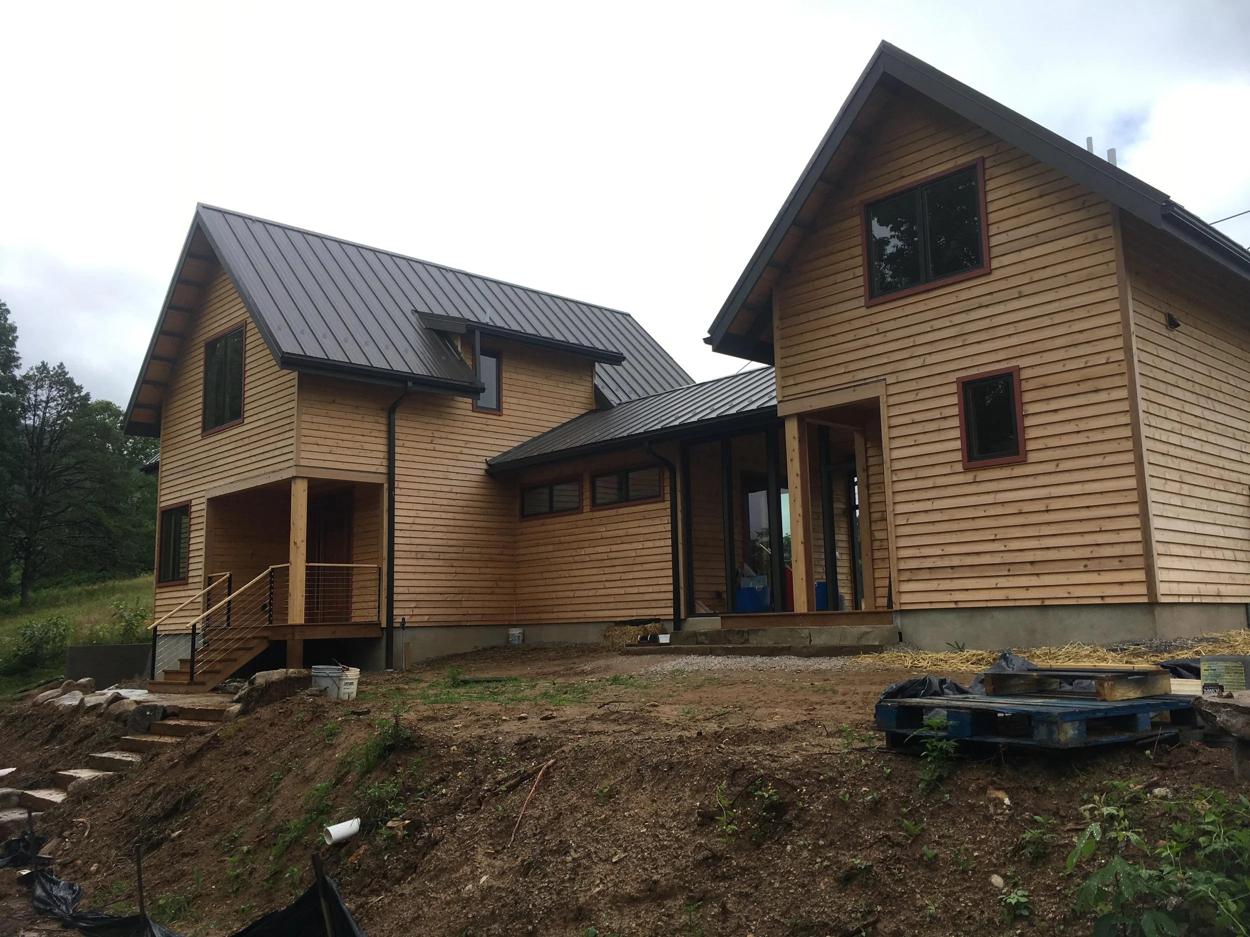 A newly constructed two-story wooden house with a metal roof, surrounded by construction materials and uneven ground, with trees in the background.